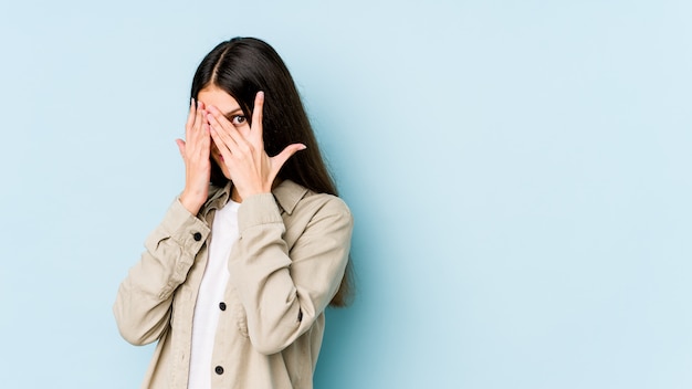 Young caucasian woman on blue wall blink through fingers frightened and nervous.