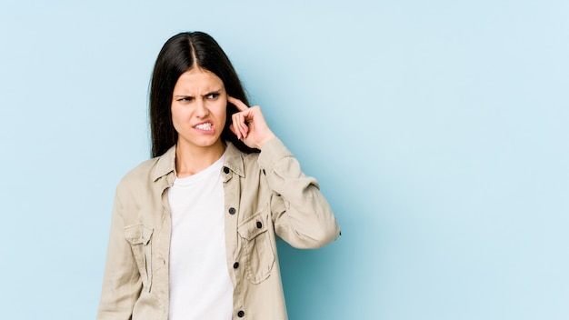 Young caucasian woman on blue wall covering ears with hands.