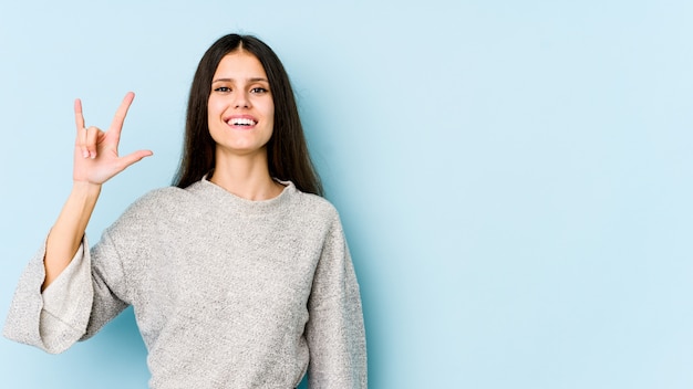 Young caucasian woman on blue wall showing a horns gesture