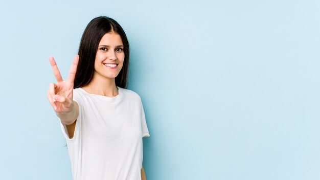 Young caucasian woman on blue wall showing victory sign and smiling broadly.