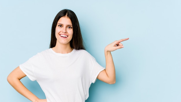 Young caucasian woman on blue wall smiling cheerfully pointing with forefinger away.