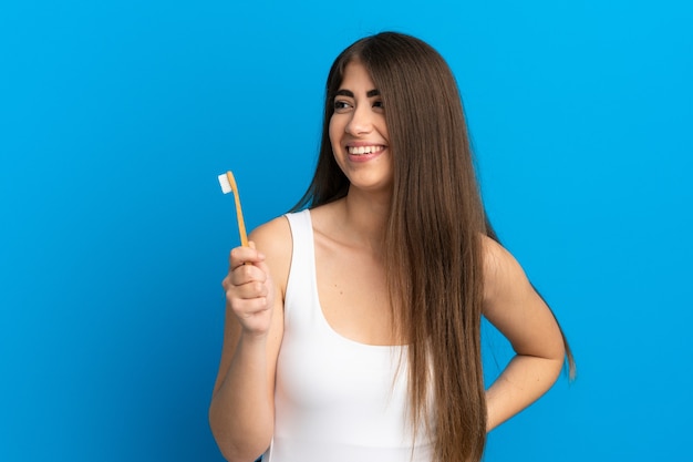 Young caucasian woman brushing teeth isolated on blue background looking to the side and smiling