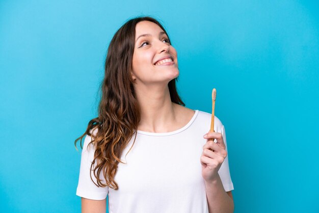 Young caucasian woman brushing teeth isolated on blue background looking up while smiling