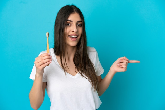 Young caucasian woman brushing teeth isolated on blue background surprised and pointing finger to the side