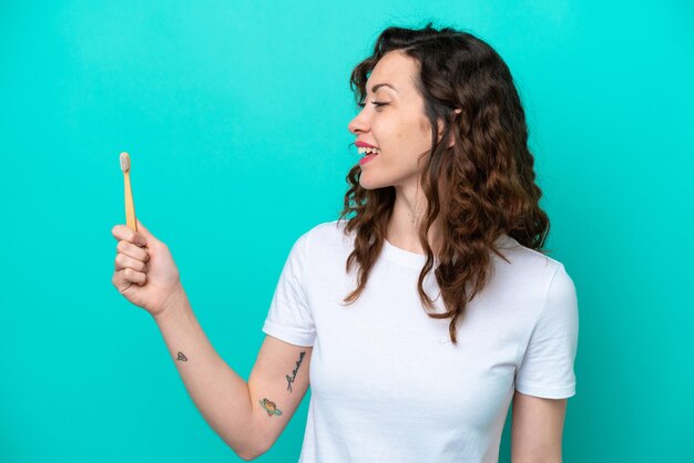 Young caucasian woman brushing teeth isolated on blue background with happy expression