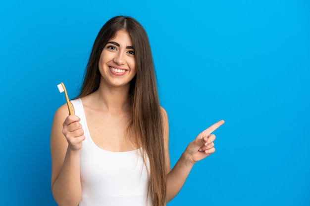 Young caucasian woman brushing teeth isolated on blue surface pointing to the side to present a product