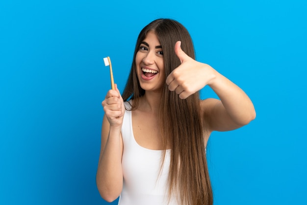 Giovane donna caucasica che si lava i denti isolati sulla superficie blu con il pollice in alto perché è successo qualcosa di buono