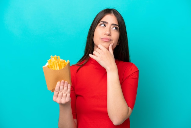 Young caucasian woman catching french fries isolated on blue background having doubts