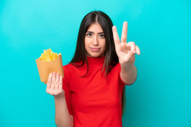 Young caucasian woman catching french fries isolated on blue background smiling and showing victory sign