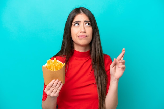 Young caucasian woman catching french fries isolated on blue background with fingers crossing and wishing the best