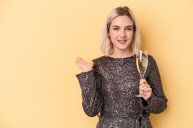 Young caucasian woman celebrating new year isolated on yellow background showing a copy space on a palm and holding another hand on waist.