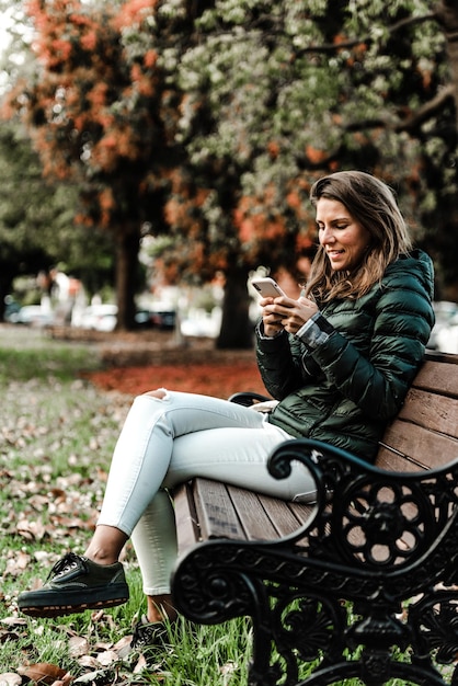 Giovane donna caucasica controlla il suo telefono nel parco durante la primavera si siede su una panchina