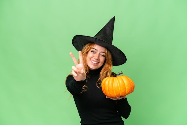 Young caucasian woman costume as witch holding a pumpkin isolated on green screen chroma key background smiling and showing victory sign