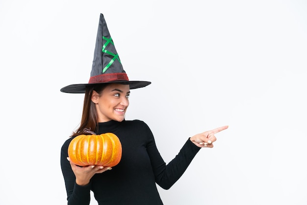 Young caucasian woman costume as witch holding a pumpkin isolated on white background pointing to the side to present a product