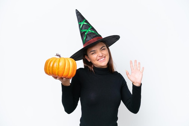 Young caucasian woman costume as witch holding a pumpkin isolated on white background saluting with hand with happy expression