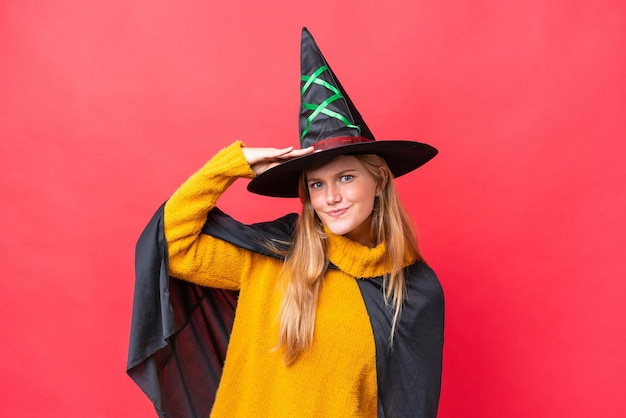 Young caucasian woman costume as witch isolated on red\
background looking far away with hand to look something