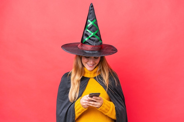 Young caucasian woman costume as witch isolated on red background sending a message with the mobile