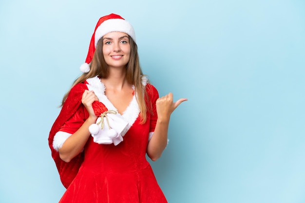 Young caucasian woman dressed as mama noel holding Christmas sack isolated on blue background pointing to the side to present a product