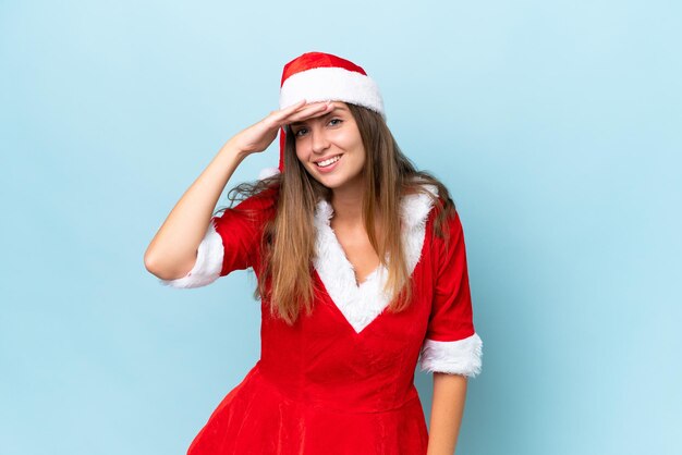 Young caucasian woman dressed as mama noel isolated on blue\
background looking far away with hand to look something