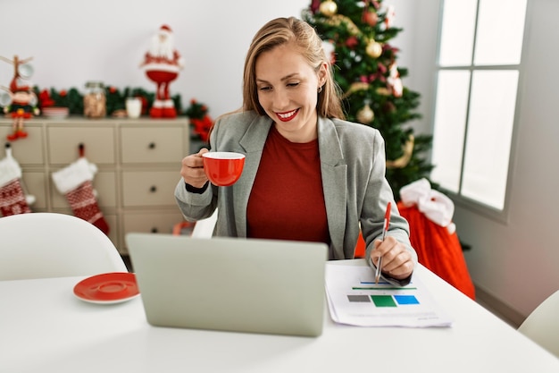 Young caucasian woman drinking coffee working sitting by christmas tree at home