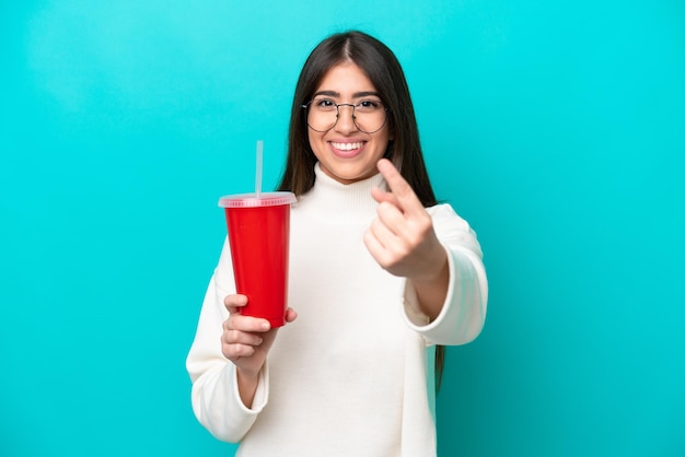 Young caucasian woman drinking soda isolated on blue background doing coming gesture