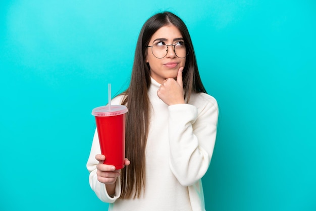 Young caucasian woman drinking soda isolated on blue background having doubts