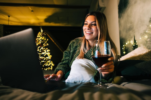 Giovane donna caucasica che beve vino davanti al computer portatile utilizzando la tecnologia in vacanza a casa sul letto.