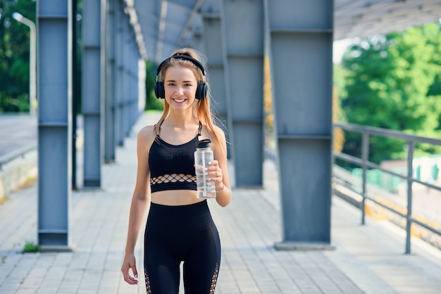 La giovane donna caucasica beve l'acqua e ascolta la musica dopo l'allenamento.