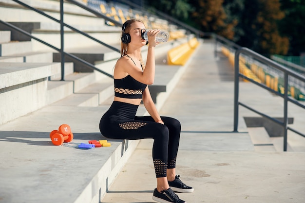 La giovane donna caucasica beve l'acqua e ascolta la musica dopo l'allenamento.