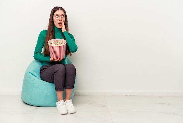 Young caucasian woman eating popcorns on a puff isolated on white background is saying a secret hot braking news and looking aside