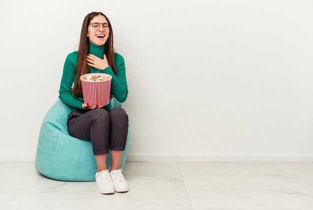 Young caucasian woman eating popcorns on a puff isolated on white background laughs out loudly keeping hand on chest.