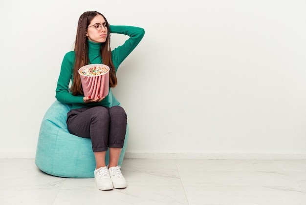 Young caucasian woman eating popcorns on a puff isolated on white background touching back of head thinking and making a choice