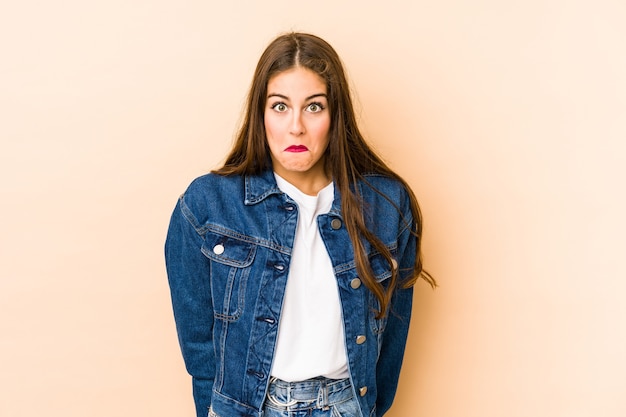 Young caucasian woman en beige shrugs shoulders and open eyes confused.