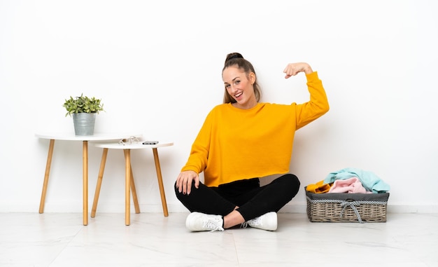 Young caucasian woman folding clothes sitting on the floor isolated on white background doing strong gesture