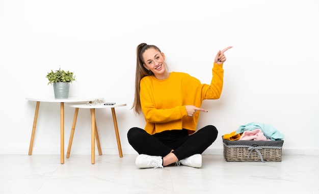 Young caucasian woman folding clothes sitting on the floor isolated on white background pointing finger to the side and presenting a product