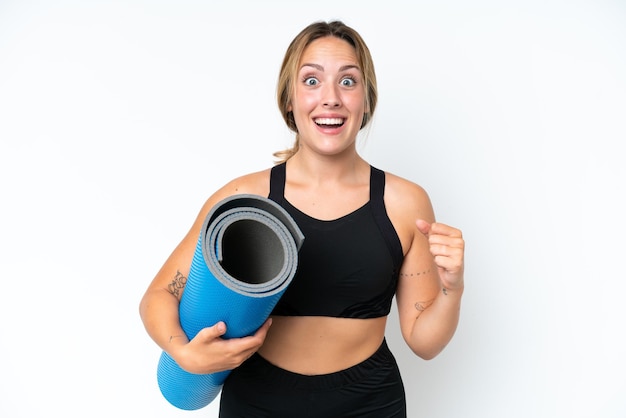 Giovane donna caucasica che va alle lezioni di yoga mentre tiene un tappetino isolato su sfondo bianco che celebra una vittoria nella posizione del vincitore