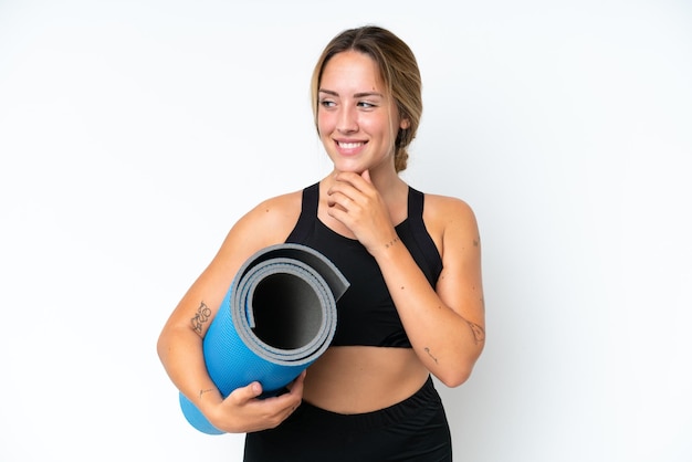 Young caucasian woman going to yoga classes while holding a mat isolated on white background looking to the side and smiling