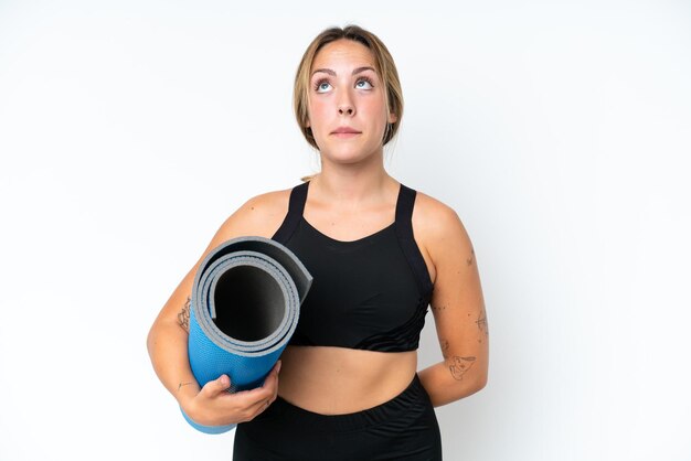 young caucasian woman going to yoga classes while holding a mat isolated on white background and looking up