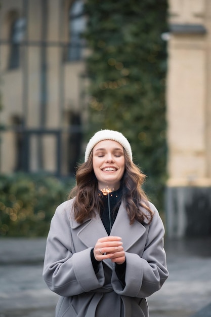 Photo young caucasian woman in gray coat with big smile holding sparkler bengal fire holiday concept