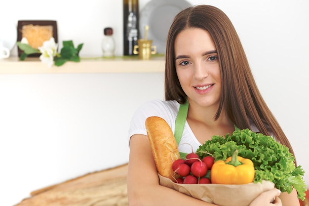 Young caucasian woman in a green apron is holding paper bag full of vegetables and fruits while smiling in kitchen.