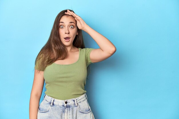 Young Caucasian woman in a green top on a blue backdrop shouts loud keeps eyes opened