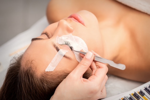 Young caucasian woman having eyelash extension procedure in beauty salon. Beautician glues eyelashes with tweezers