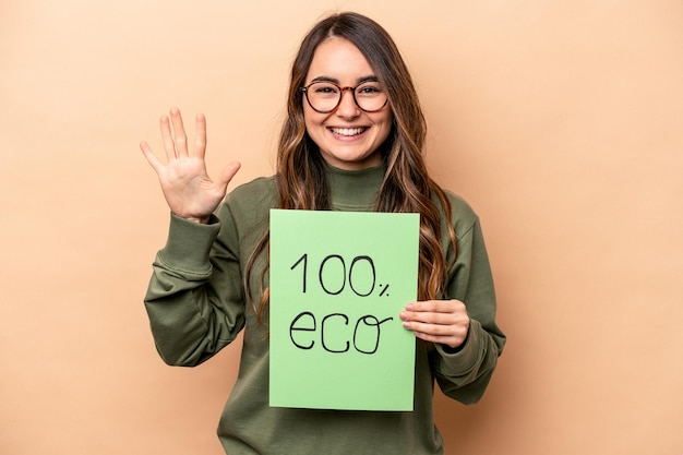 Young caucasian woman holding 100 eco placard isolated on beige background smiling cheerful showing number five with fingers