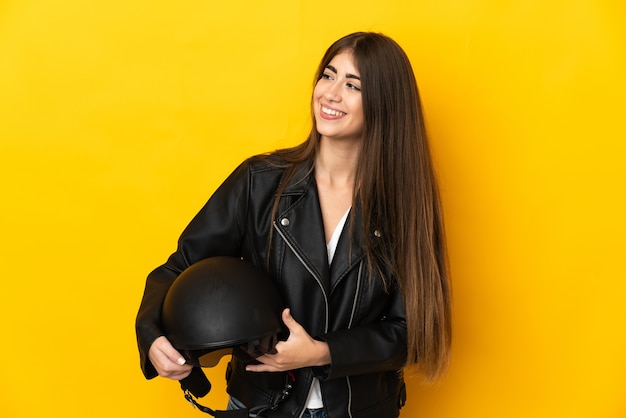 Молодая кавказская женщина, держащая мотоциклетный шлем, изолирована на желтом фоне, смотрит в сторону и улыбается