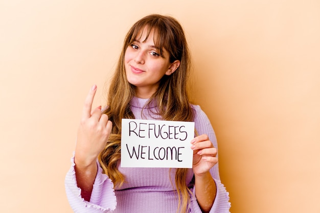 Фото Молодая кавказская женщина, держащая приветственный плакат беженцев, изолирована, указывая пальцем на вас, как будто приглашая подойти ближе.