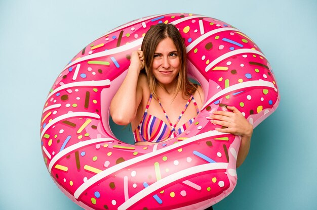 Young caucasian woman holding air mattress isolated on blue background covering ears with hands