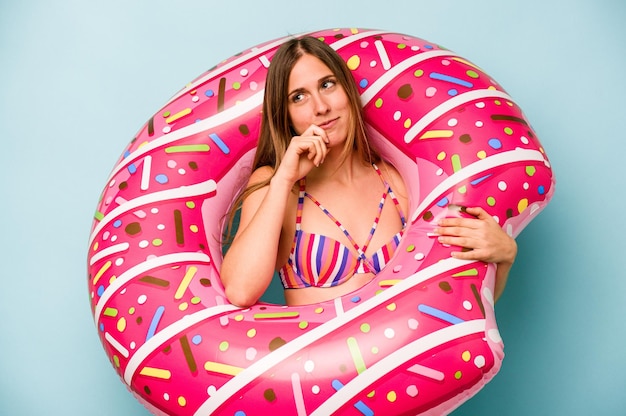 Young caucasian woman holding air mattress isolated on blue background looking sideways with doubtful and skeptical expression