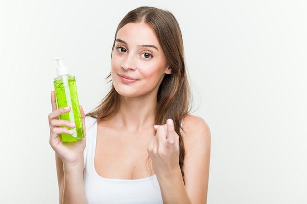 Young caucasian woman holding an aloe vera bottle pointing with finger at you as if inviting come closer.