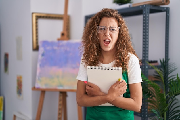 Young caucasian woman holding art notebook angry and mad screaming frustrated and furious, shouting with anger. rage and aggressive concept.