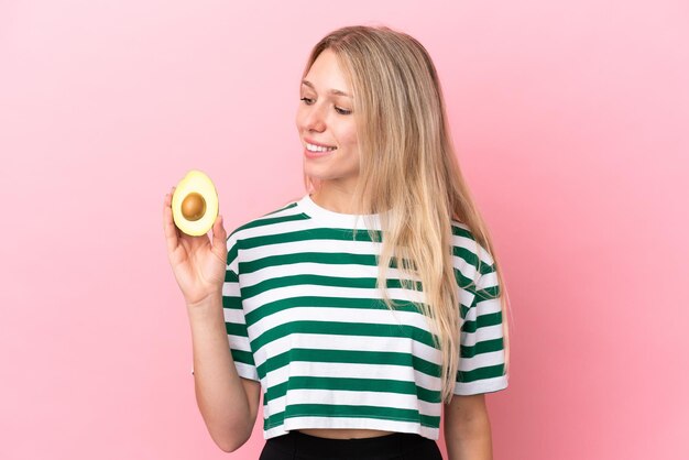 Giovane donna caucasica che tiene un avocado isolato su sfondo rosa guardando il lato